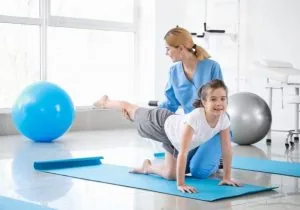 Physical therapist guiding child in plank position on exercise mat with stability balls in bright clinical room